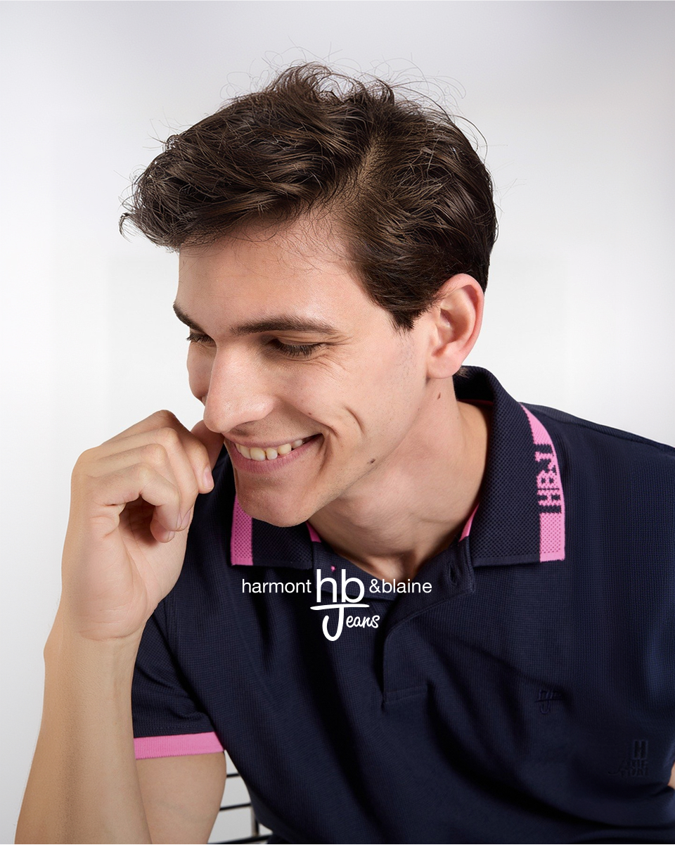 L'immagine ritrae il primo piano di un giovane uomo sorridente con capelli castani e mossi, orientato di trequarti verso sinistra mentre appoggia delicatamente il pugno alla guancia. Indossa una polo blu scuro con dettagli rosa sul colletto e sui bordi delle maniche, completata dalla scritta rosa "HBJ" sul colletto e dal logo sovrapposto "harmont & blaine hb Jeans" al centro.
